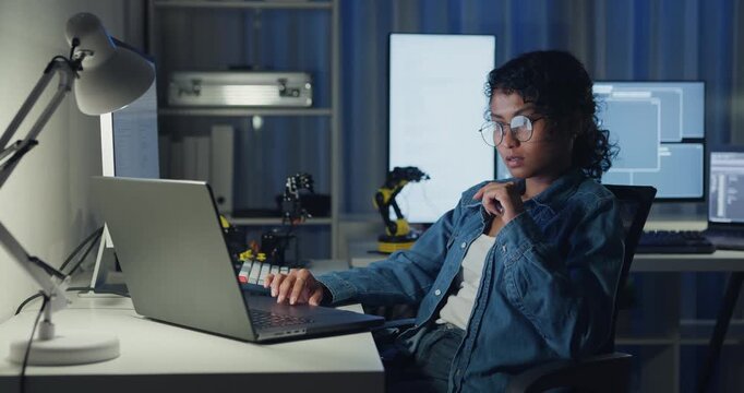 Young African American female programmer coding AI software on multiple screens in night office. Network security, cybersecurity, machine learning, working late, overtime, robotics automation concept.