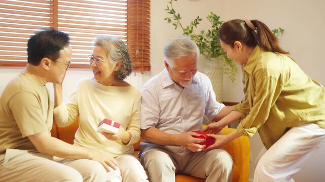 Happy Asian adult child giving gifts to senior parents at home. Multi-generation family sharing joyful surprise moment. Grandparents and family members enjoying bonding and love across generations