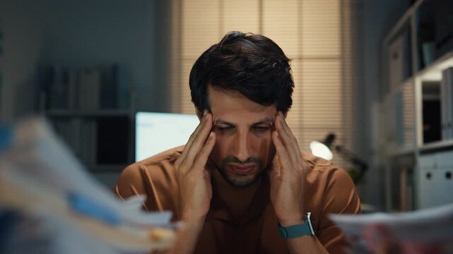 Stressed young Latin businessman having severe headache and migraine at desk in night office. Debt crisis, corporate burnout, late overtime workload, mental health anxiety, business failure concept.