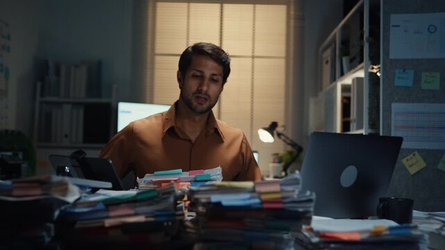 Stressed young Latin businessman walking to place tax document stack at desk in dark night office. Overworked overtime, heavy workload stress, tired employee, corporate business burnout concept.