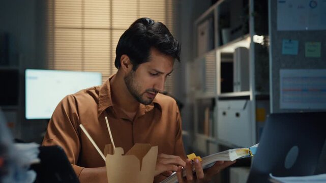 Young Latin businessman eating takeout instant noodles and reviewing document on laptop at desk in dark night office. Overtime working late, overworked deadline, fast food, corporate problem concept.
