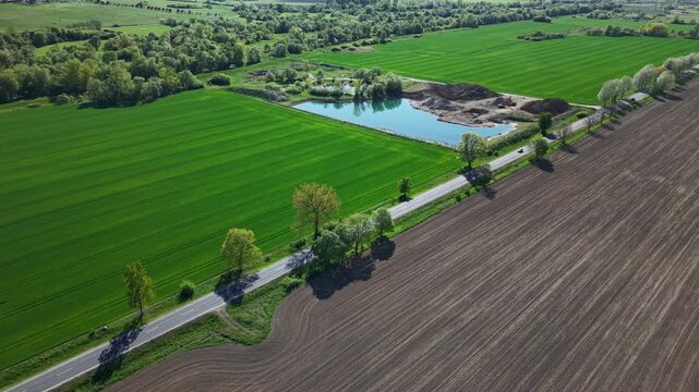 An aerial perspective shows a road dissecting vibrant green fields and ploughed soil with a small blue pond nearby.