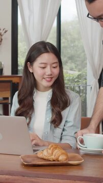 Vertical Screen : Young Asian freelance women work talk on phone with customer at coffee shop. Asian happy men barista waiter wear gray apon serve hot coffee to female customer who business in cafe.
