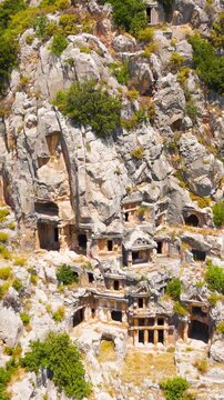 Vertical video. Demre, Turkey. Ancient Lycian rock-cut tombs with temple-style facades carved into a sheer limestone cliff face, close-up aerial drone view in bright summer daylight. Aerial View. Ric