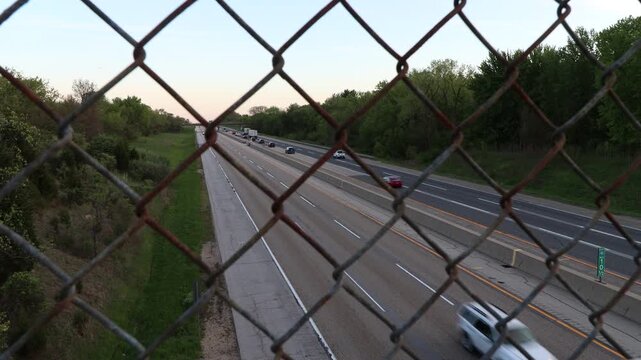 A metal rusted chain link fence out of focus with cars and semi trucks driving down the highway road behind the fence. 