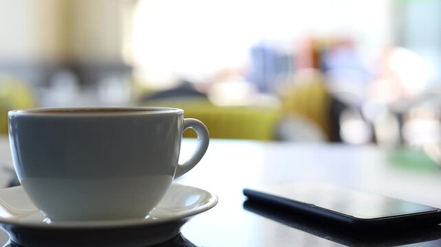 Close-up of a white coffee cup and smartphone placed on a table in a modern cafe with blurred background and soft natural light. Cozy lifestyle, morning routine, remote work and relaxation concept.