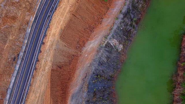 Vertical drone perspective showcasing striking geological contrasts. Vibrant red soil, grey rocky terrain, and emerald water sit adjacent to a modern asphalt road. Industrial and natural textures comb