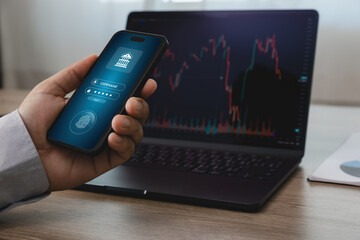 Hand holding smartphone with mobile online banking login screen in front of laptop displaying...