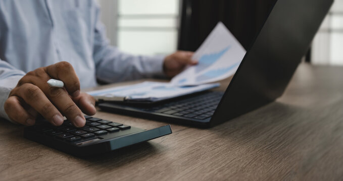 Business person using calculator with financial documents and laptop on office desk, suitable for finance accounting, investment analysis, budgeting, expense calculation, and financial reporting.