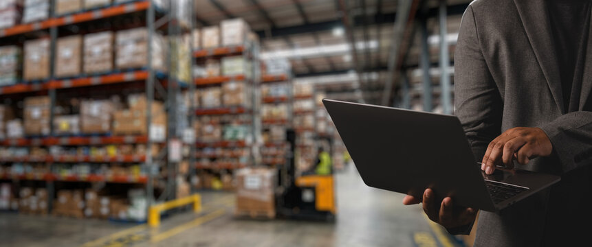 Worker using laptop in modern warehouse with storage shelves and cargo boxes, suitable for smart warehouse management system, logistics monitoring, inventory tracking, and industrial supply operations