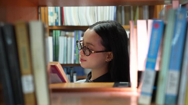 Asian woman student reading a book in the library. Female college student wearing glasses and smiling while studying near bookshelves in university. looking through a stack of books in school .