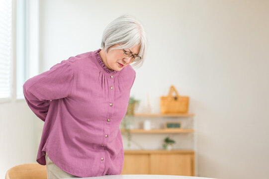 A senior woman with gray hair, clutching her lower back in apparent pain.