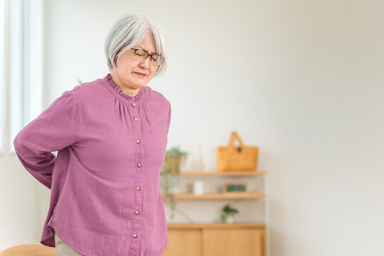 A senior woman with gray hair, clutching her lower back in apparent pain.