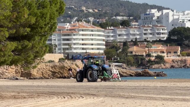 Palma, spain, February 18, 2026, tractor cleaning and raking a sandy beach to prepare for summer tourist season with modern hotels in the background