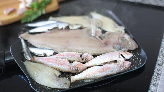 Carcasses of raw Atlantic fish of various species are laid out in pan. Process of cooking megrim, European anchovy, striped mullus barbatus on induction cooker. Assorted fish, meagre diet sea fish