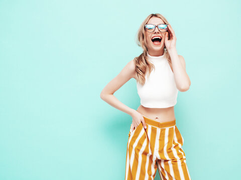 Young beautiful smiling blond female in trendy summer yellow clothes. Sexy carefree woman posing near blue wall in studio. Positive model having fun indoors. Cheerful and happy. In spectacles