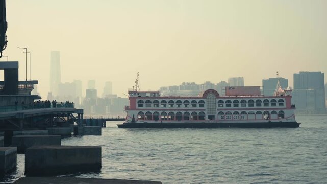 Static wide shot of a double-deck passenger ferry arriving at a waterfront pier in an Asian coastal city, with dense hazy skyline and mountains visible in background, calm and atmospheric daytime scen