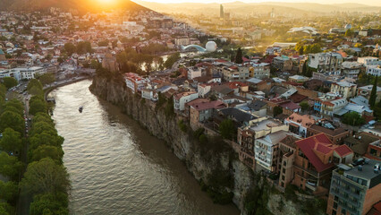 Aerial sunset view of Metekhi district in Tbilisi, Georgia, featuring historic houses on the Kura River cliffs with warm golden hour light over the city © Michele
