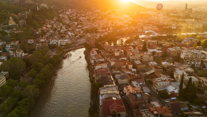 Aerial sunset view of the Metekhi district in Tbilisi, Georgia, with historic houses perched on high cliffs above the Kura River under a warm golden glow © Michele