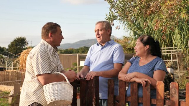 Couple of smiling elderly amateur gardeners discussing interesting topics with their male neighbor while standing near wooden fence in garden on summer day