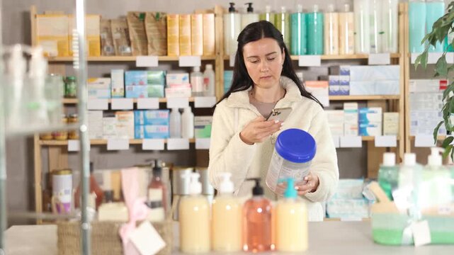 Adult woman stands in a pharmacy near the shelves with goods and scans the barcode of a jar of baby food. Buyer pays for baby food using a QR code in the mobile application. High quality 4k footage