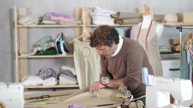  Adult male tailor cutting out pattern from paper with scissors in sewing workshop