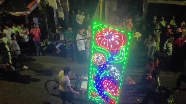HOWRAH, INDIA - MAY 11, 2026: Cinematic nighttime footage capturing the energetic atmosphere of a local street festival. A dense crowd gathers to watch an intricate, animated LED light procession feat