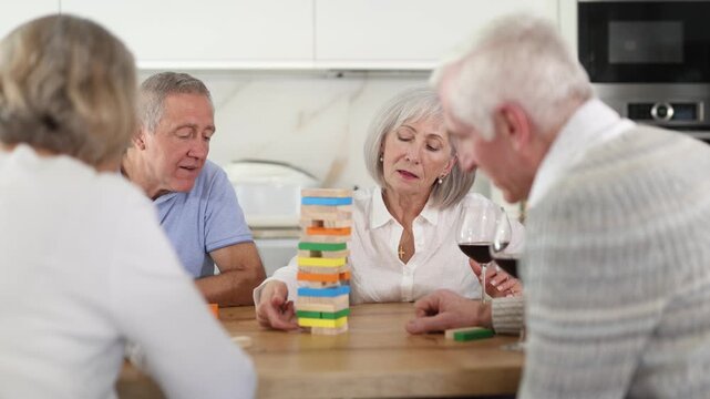 Group of four mature friends women and men is playing board game. Two senior married couples build tower with wooden blocks bricks 