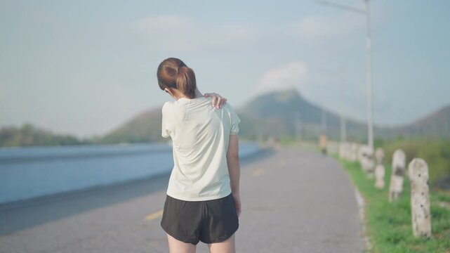 Young Asian fitness woman holding her injured shoulder and neck due to muscle pain during training. Runner female having body problem after outdoor exercise in summer. Sports and medical concept