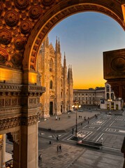 Fototapeta premium Golden sunset light frames the majestic Gothic cathedral through ornate arches in Milan, creating a timeless architectural scene filled with elegance, symmetry, and historic European charm.
