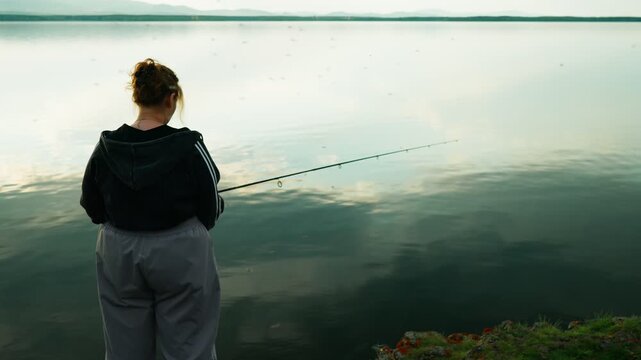 A female fisherman is fishing on a lake, surrounded by a swarm of blood-sucking mosquitoes that are causing her discomfort. Real footage with no staged elements.