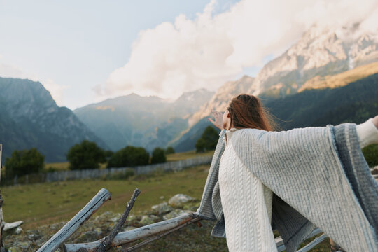 Young woman with curly hair wrapped in a cozy knitted shawl enjoys fresh mountain air while standing near a wooden fence in a serene rural landscape.
