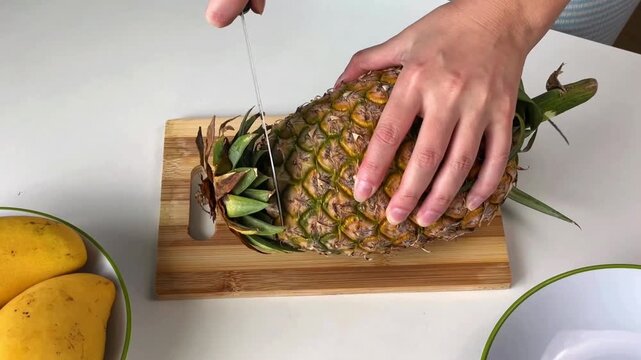 Women's hands peel a fresh pineapple and cut off the top of the fruit with a knife on a wooden board.