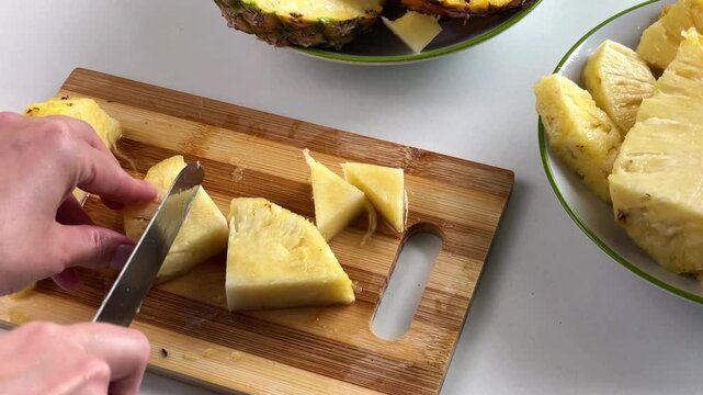 A woman's hands slice a fresh pineapple into pieces and trim away the tough, inedible part of the fruit on a wooden cutting board.