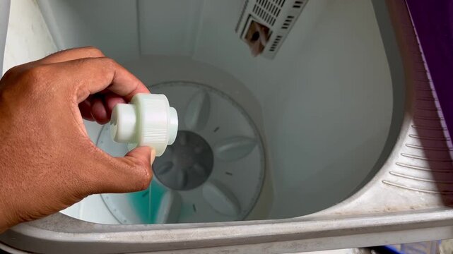 Close-up of a person pouring blue liquid detergent into a top-loading washing machine.