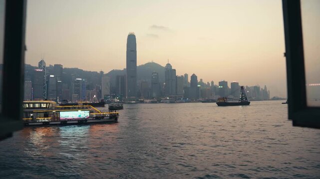 Static framed shot through window of an illuminated cruise boat sailing across Victoria Harbour at dusk, with Hong Kong island skyline, IFC tower, and Victoria Peak visible, cinematic and atmospheric