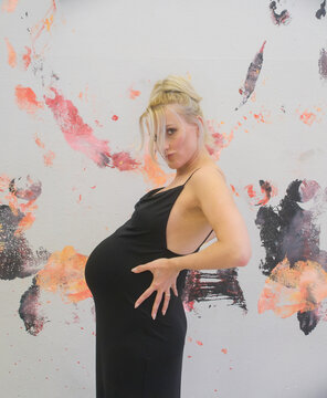 Pregnant woman in black dress posing in front of expressive abstract paint wall strong gesture and confident posture highlight belly creative artistic maternity portrait