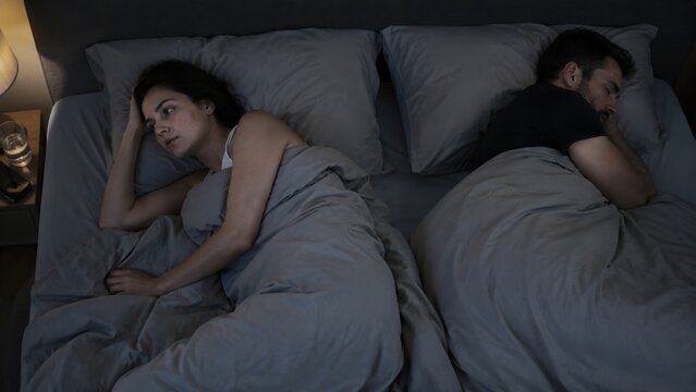 Awake adult disturbed by restless sleep beside calm sleeping partner, neutral bedding, modern bedroom scene about insomnia and relationship sleep problems