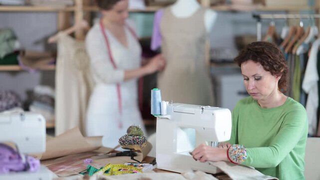 Female dressmaker sewing at machine while colleague working with mannequin