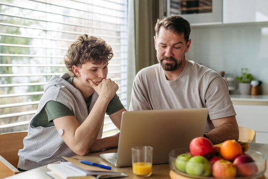 Father helping teenage son with diabetes with homework on laptop in kitchen.