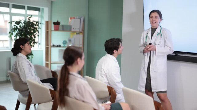  Young female doctor in medical gown with remote control gives presentation to colleagues doctors in conference room
