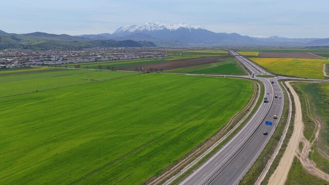 Highway A3 and roundabout in Cristian village with snow capped Bucegi mountains, scenic spring landscape and green farmland, aerial ascending view and panoramic pan, 4K Cinematic