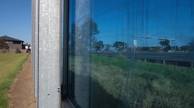 Transparent noise barrier wall alongside a highway in Melbourne,  Australia, with busy traffic visible in the panel, near a modern residential home, green public space. Infrastructure to reduce noise
