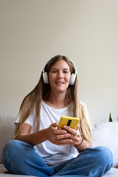 Young woman listening music with headphones and using smartphone on sofa at home