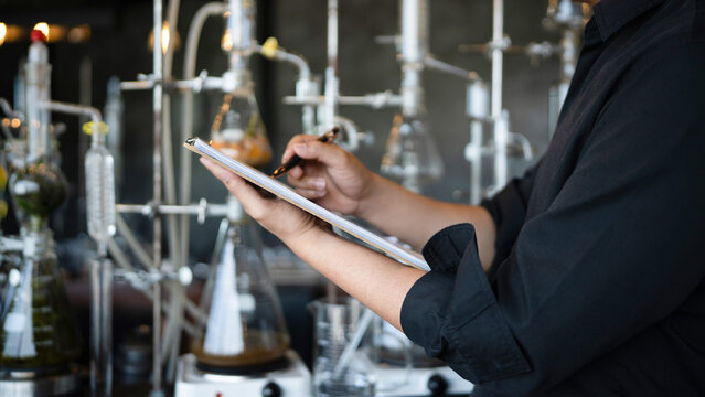 A researcher in a black lab coat writes observations on a clipboard surrounded by glass distillation apparatus, ideal for science and pharmaceutical concepts