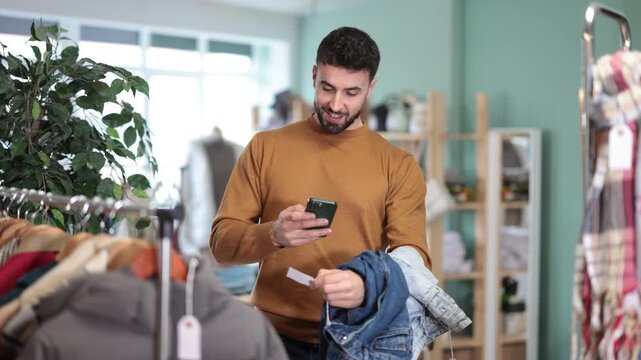 Bearded man scans on mobile camera qr on jeans labels. Man pays for clothes with a modern payment method through a mobile application. High quality 4k footage