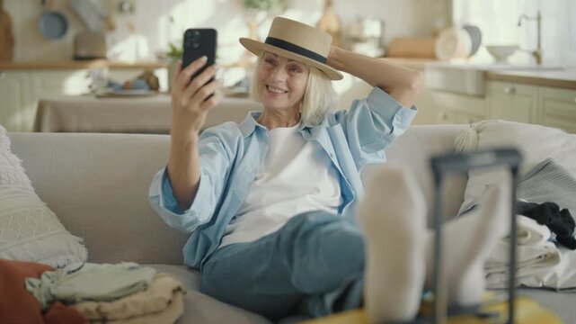 Emotional woman sharing her expressions and emotions of summer tourist trip. Mature lady talking by video call in smartphone, laughing and smiling when sitting in living room apartment, good mood