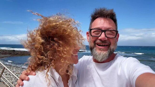 Cheerful middle-aged couple having fun and enjoying summer vacation while filming a selfie video together. The woman's curly hair is blowing in the wind as they smile and laugh at the camera