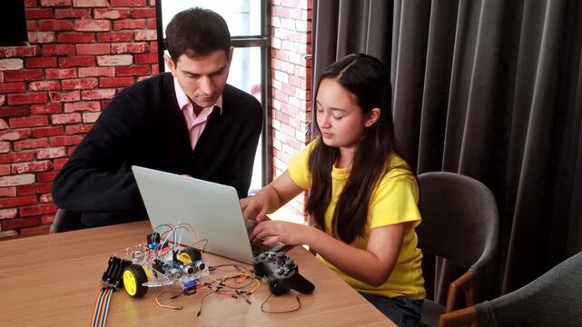 A student and mentor work on a laptop beside a small wheeled robot with wires and a microcontroller, building and testing a STEM electronics project in a modern classroom.