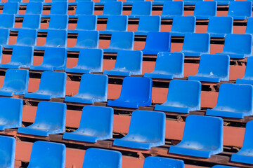 Fototapeta premium Empty blue plastic stadium seats are arranged in rows on a wooden bleacher structure, illuminated by bright sunlight in an outdoor setting